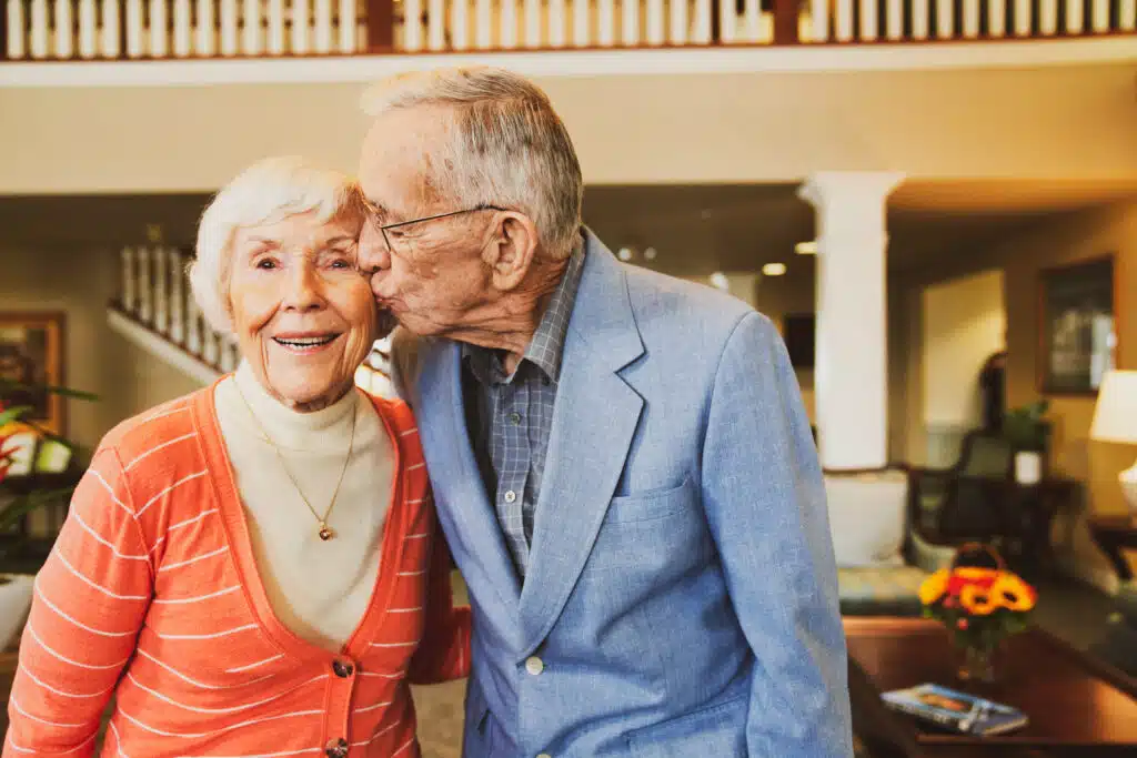 Elderly man kisses smiling woman in cozy room.