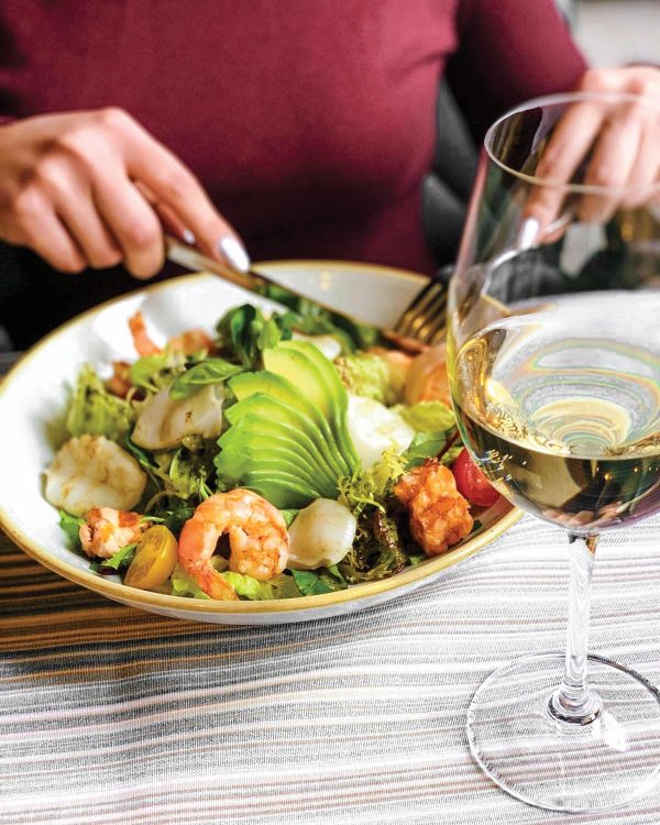 A close-up of someone eating a large salad with shrimp and avocado, with a glass of wine to the side