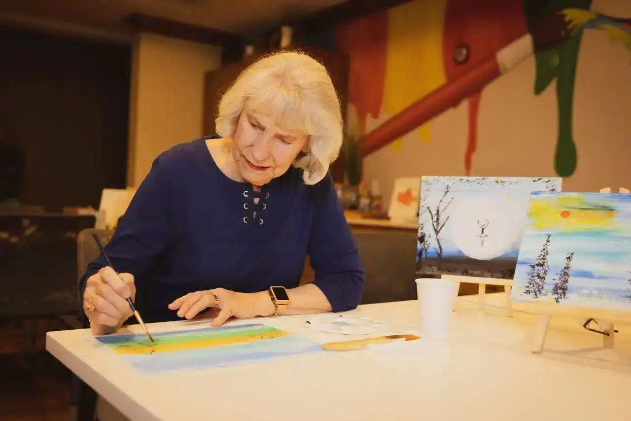 Elderly woman painting at a table, colorful surroundings.